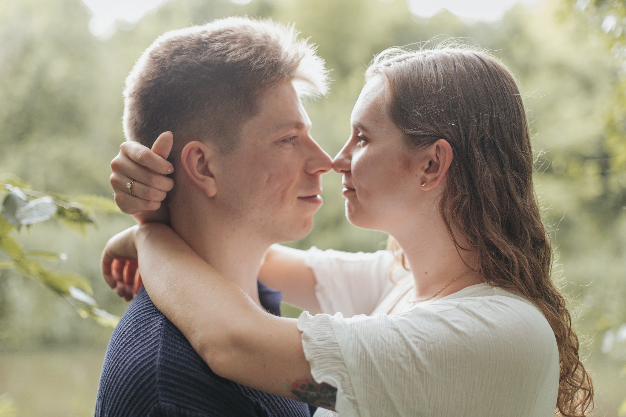 Zakochana para, zbliżenie twarzy, dotykają się nosami. Ona obejmuje go rękami, widać pierścionek zaręczynowy. Tło rozmyte, zielone. Romantyczna sesja narzeczeńska.
