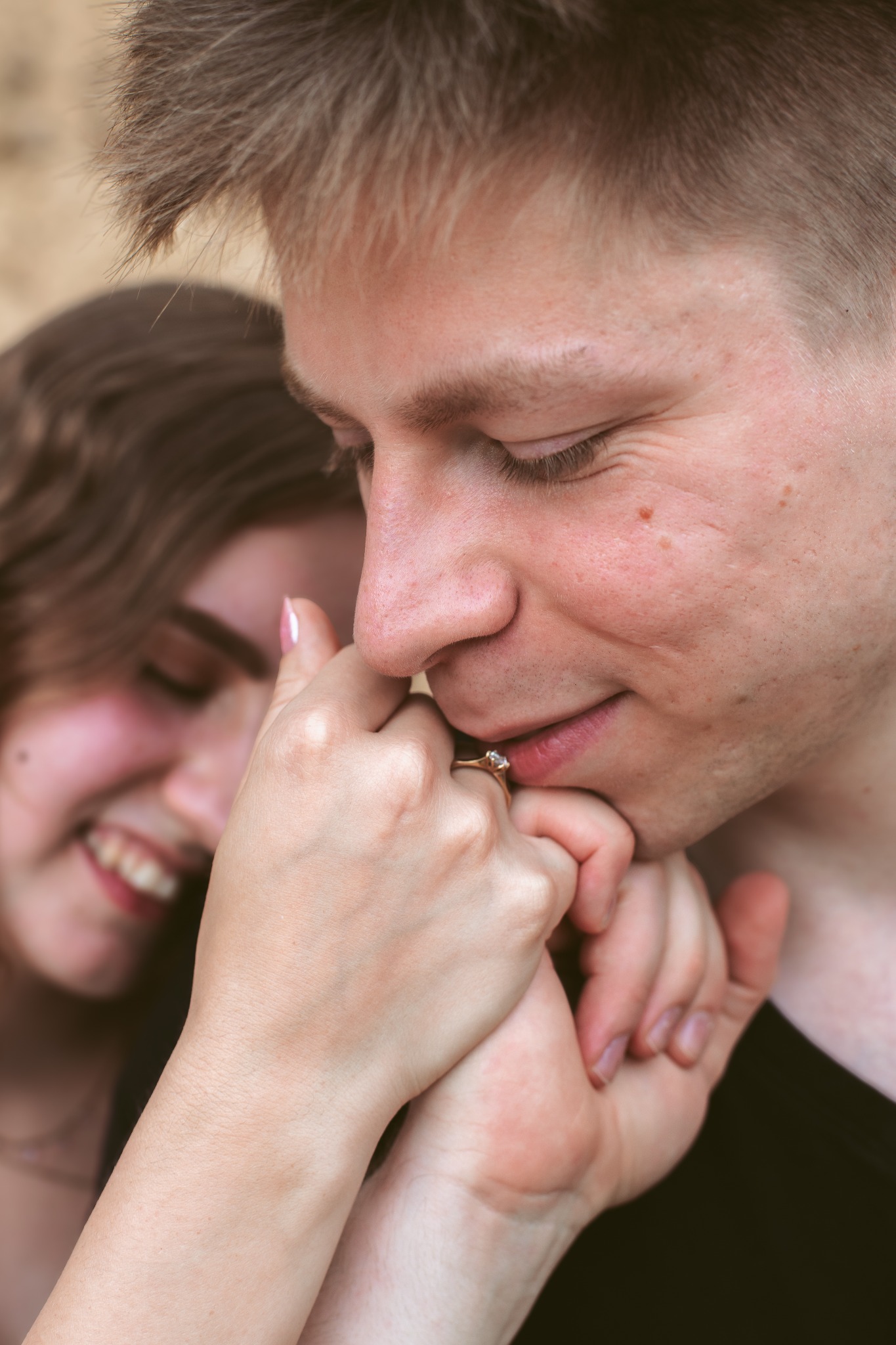 Zakochana para: mężczyzna delikatnie całuje dłoń kobiety z pierścionkiem zaręczynowym, ona uśmiecha się w tle, intymna chwila, ciepłe oświetlenie.