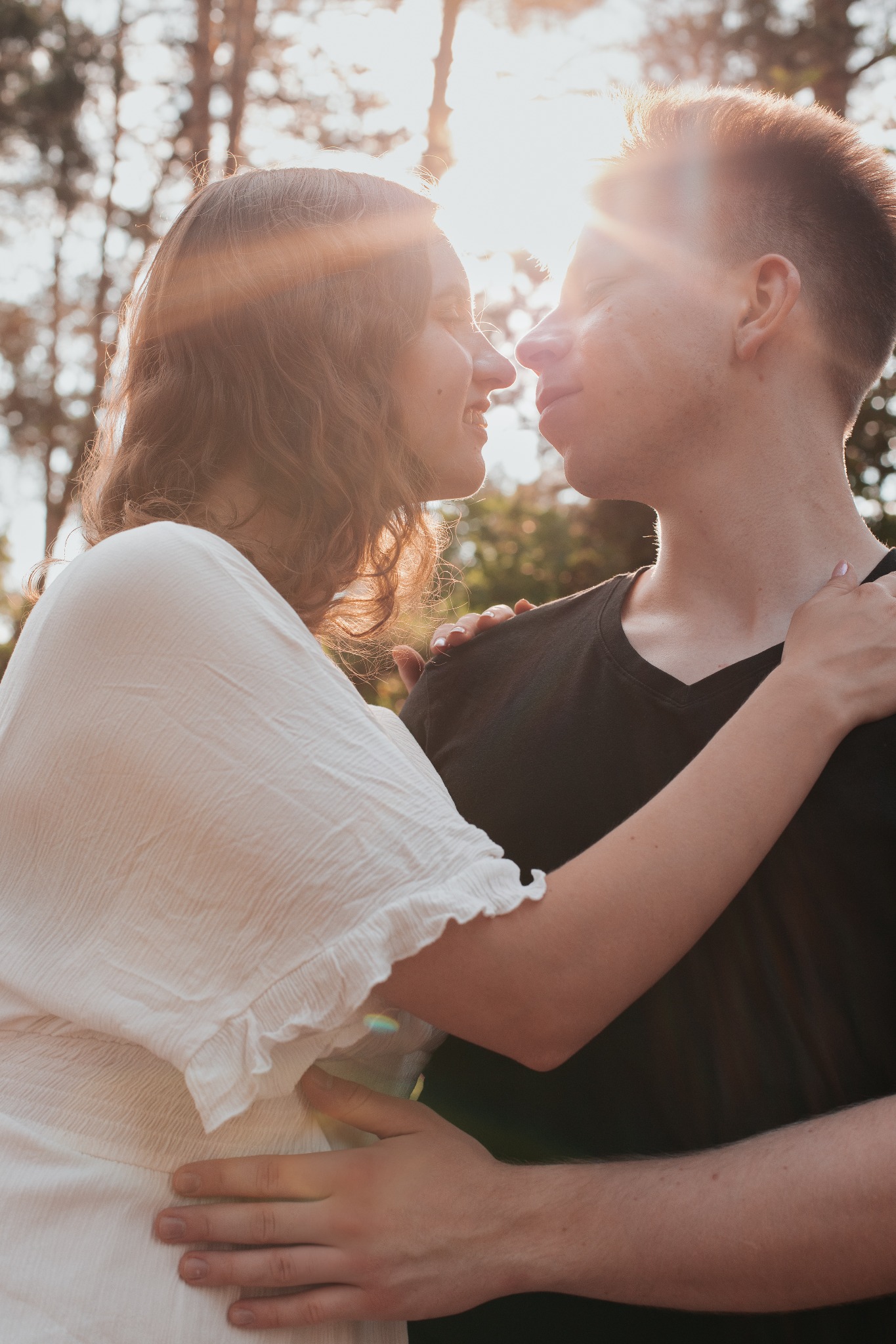 Młoda para w objęciach, zbliżająca się do pocałunku w słonecznym lesie. Dziewczyna w białej bluzce, chłopak w czarnej koszulce. Romantyczna atmosfera.