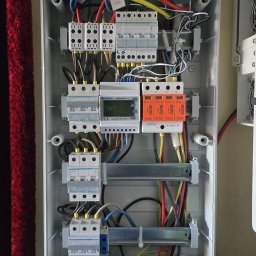 PV-EL - Otwarta skrzynka elektryczna z bezpiecznikami, przewodami i licznikiem energii. Widoczne elementy instalacji fotowoltaicznej.