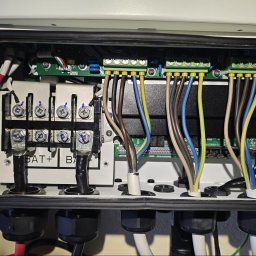 PV-EL - Wnętrze skrzynki elektrycznej z podłączeniami akumulatorów i przewodami o różnych kolorach. Widoczne złącza, śruby i elementy elektroniczne na płytkach drukowanych.