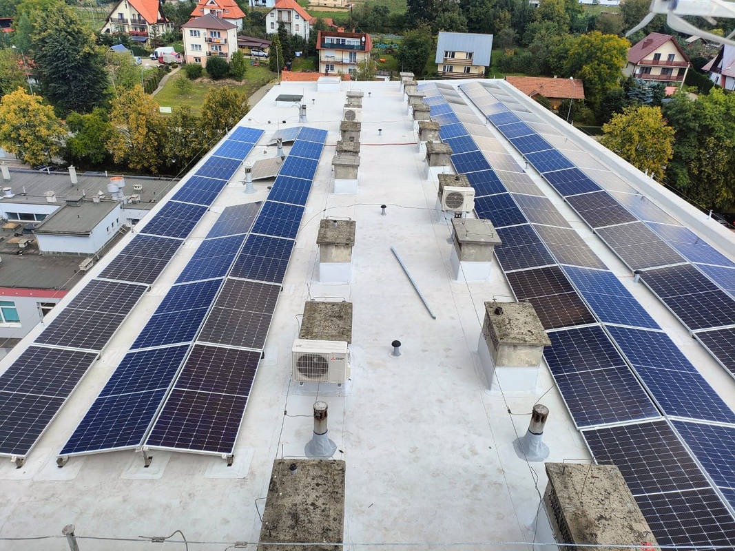 Widok z góry na instalację paneli słonecznych na płaskim dachu budynku z kominami i klimatyzatorami, w tle domy jednorodzinne i drzewa.