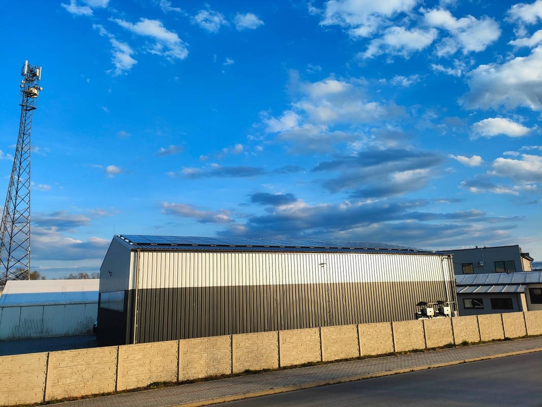 Metalowy budynek z panelami słonecznymi na dachu, widoczna wieża telekomunikacyjna w tle, błękitne niebo z chmurami.