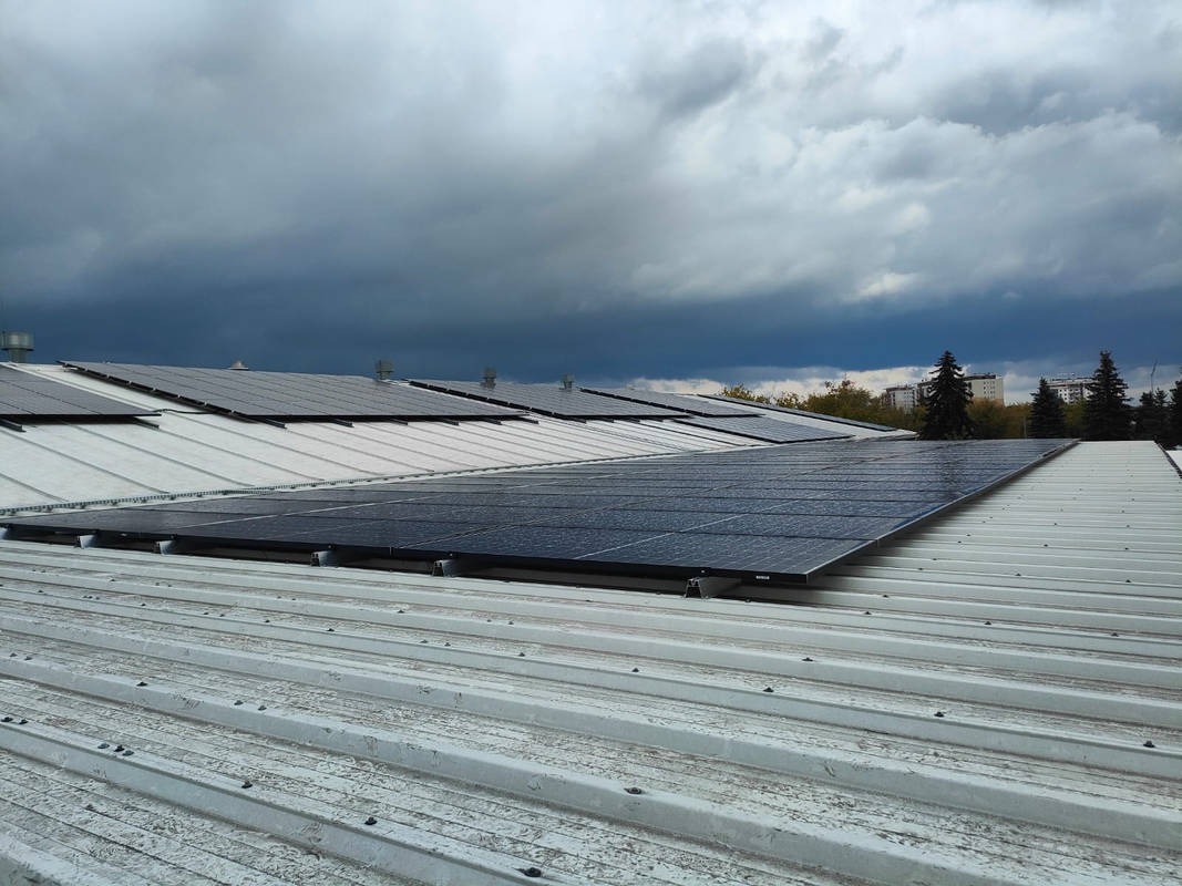 Rzędy paneli słonecznych na blaszanym dachu budynku przemysłowego, widoczne krople deszczu na panelach, pochmurne niebo w tle.