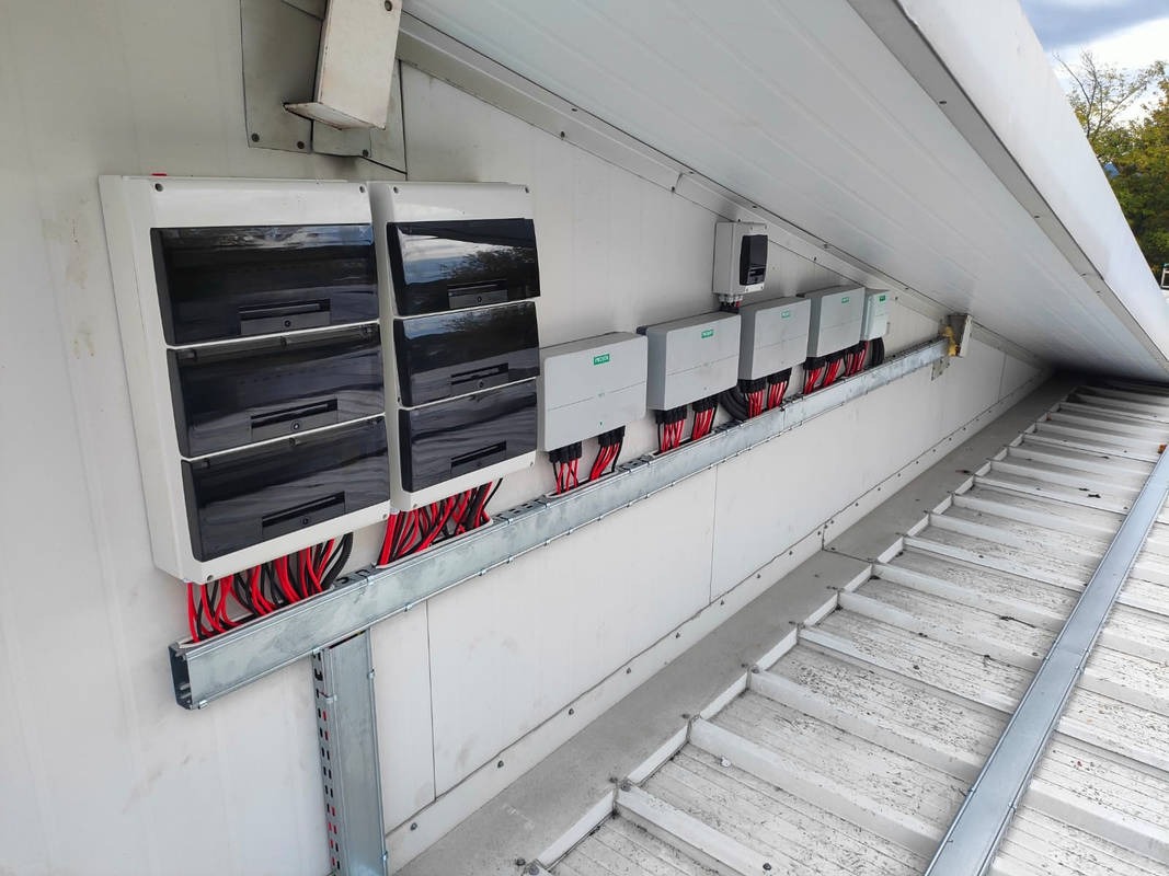 Szereg falowników i rozdzielnic połączonych przewodami na metalowej szynie montażowej, instalacja na zewnątrz budynku z blachy trapezowej.