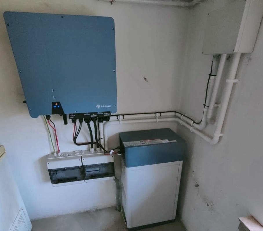 Instalacja fotowoltaiczna w pomieszczeniu: falownik marki Solplanet, skrzynka zabezpieczeń i magazyn energii w obudowie dwukolorowej, biało-niebieskiej, połączone rurkami i przewodami.