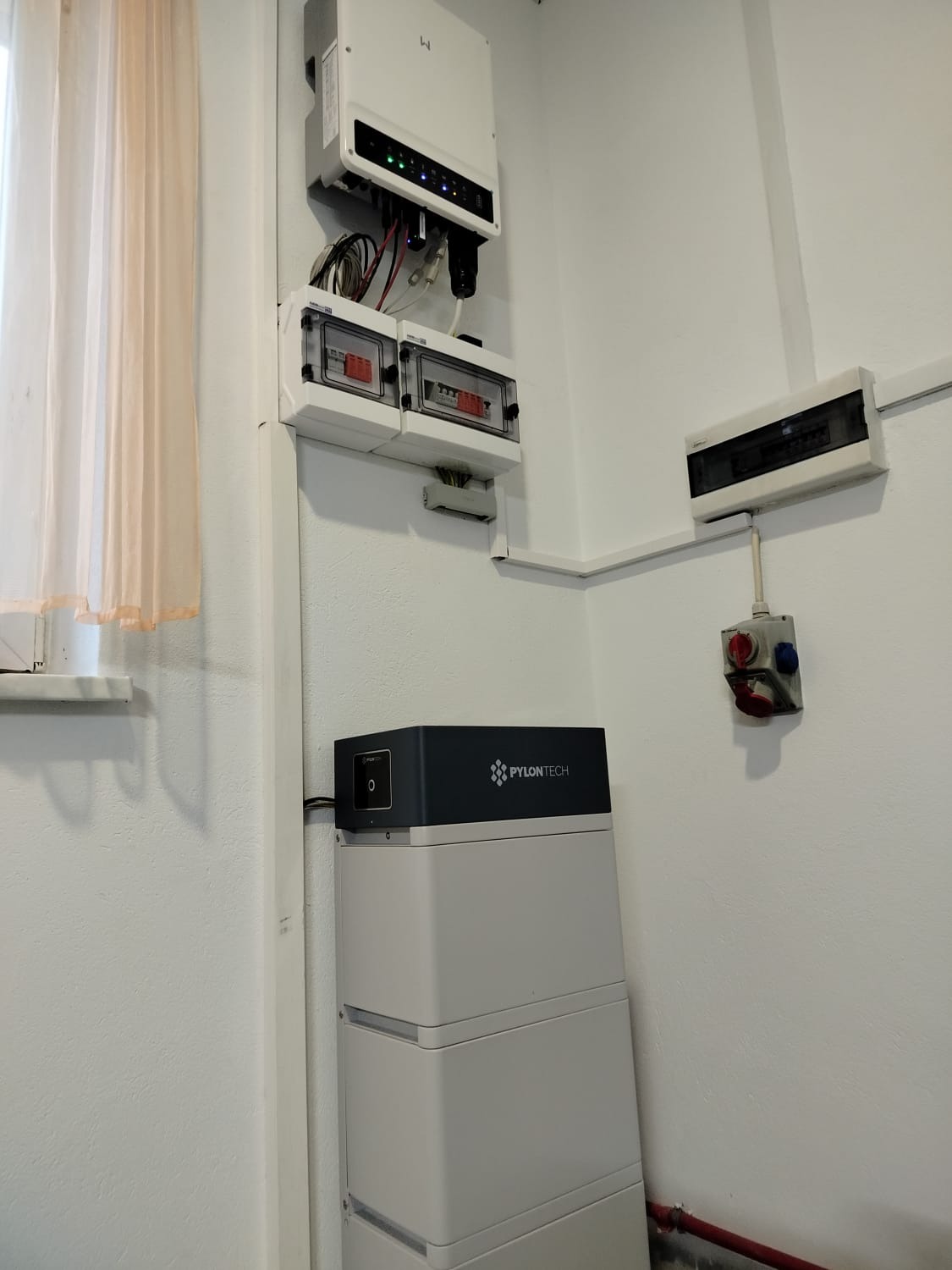 Bateria magazynu energii Pylontech ułożona z trzech modułów, obok okna z zasłoną, na ścianie inwerter z okablowaniem i skrzynki bezpiecznikowe.