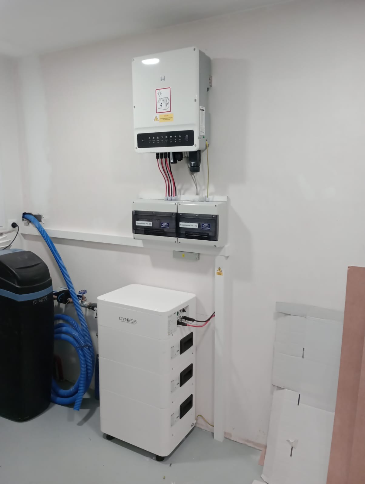 Biały magazyn energii Dyness, falownik fotowoltaiczny na ścianie, zabezpieczenia elektryczne i zmiękczacz wody w pomieszczeniu technicznym z szarą podłogą.