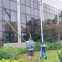 Pracownik w szarym t-shircie myje okna na wysokości w budynku biurowym za pomocą długiej lancy z niebieską szczotką, stojąc na trawniku obok wózka z niebieskimi zbiornikami na wodę.