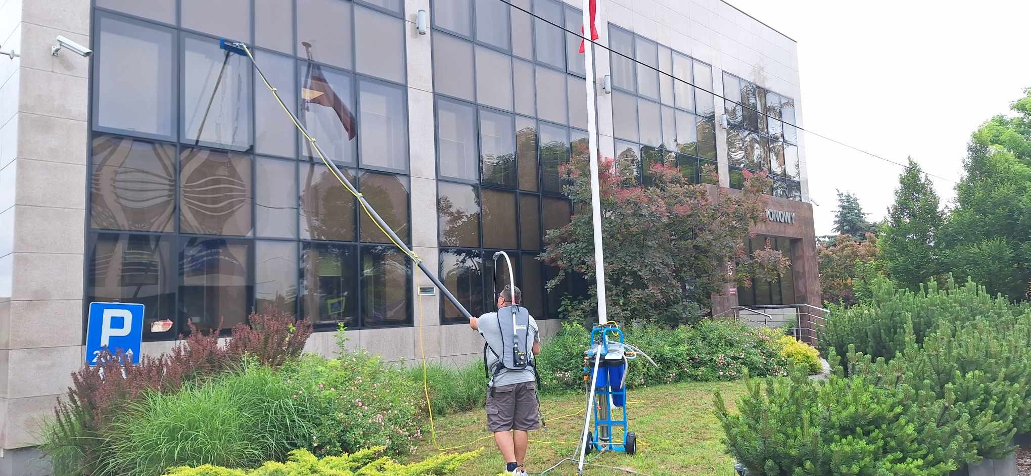 Pracownik w szarym t-shircie myje okna na wysokości w budynku biurowym za pomocą długiej lancy z niebieską szczotką, stojąc na trawniku obok wózka z niebieskimi zbiornikami na wodę.