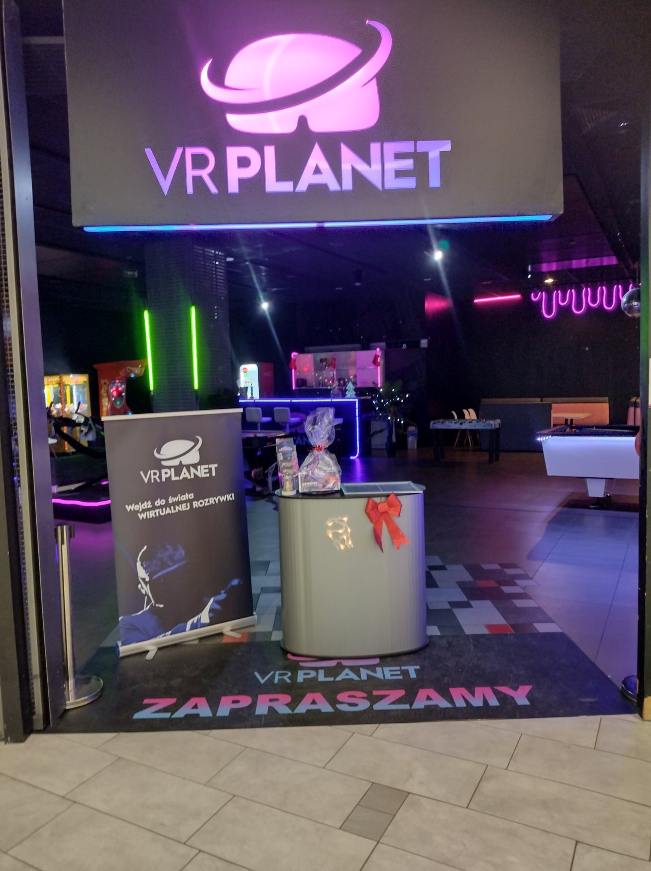 Wejście do salonu VR Planet z logo, roll-upem reklamowym i stanowiskiem recepcyjnym z prezentem. W tle widoczne stanowiska do gier VR i neonowe oświetlenie.