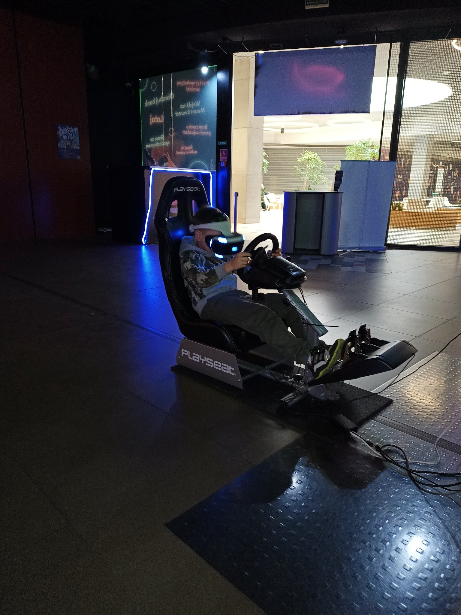 Chłopiec w okularach VR siedzi na symulatorze wyścigowym Playseat z kierownicą i pedałami, w tle widać jasne wnętrze centrum handlowego.