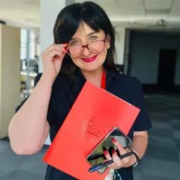 Uśmiechnięta kobieta w okularach, trzymająca czerwony folder z logo Generali, smartfon i długopis w biurze.