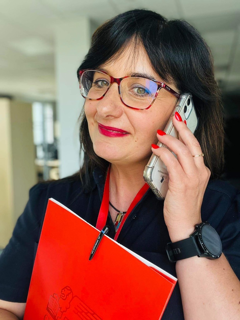 Uśmiechnięta kobieta w okularach rozmawia przez telefon, trzymając czerwony segregator z wytłoczonym logo i czarnym długopisem wpiętym w okładkę.