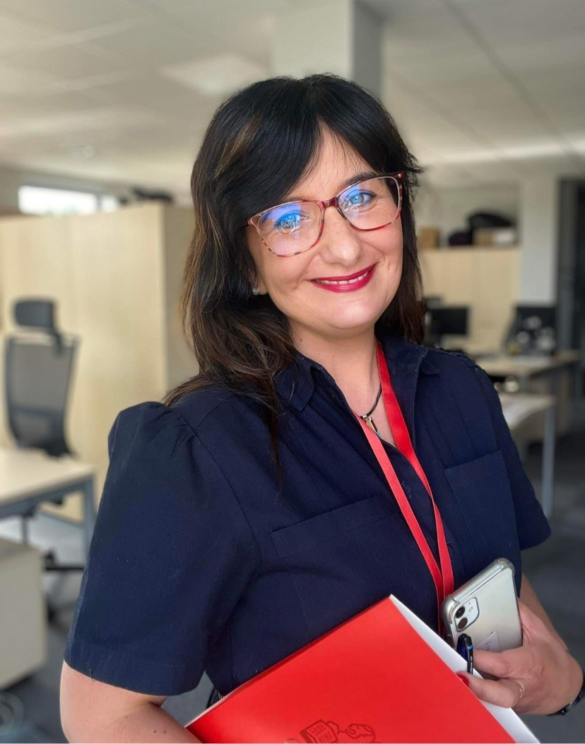 Uśmiechnięta kobieta w okularach, ubrana w granatową koszulę, trzymająca czerwony segregator i telefon komórkowy w biurze z jasnym oświetleniem.
