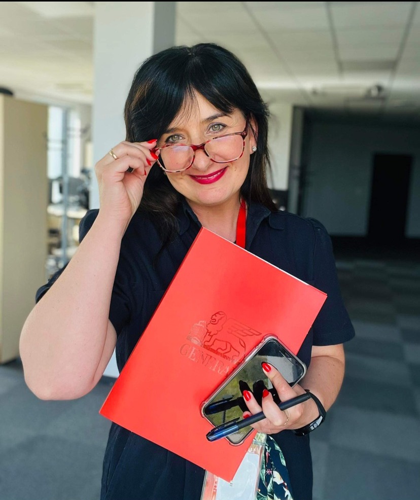 Uśmiechnięta kobieta w okularach, trzymająca czerwony folder z logo Generali, smartfon i długopis w biurze.