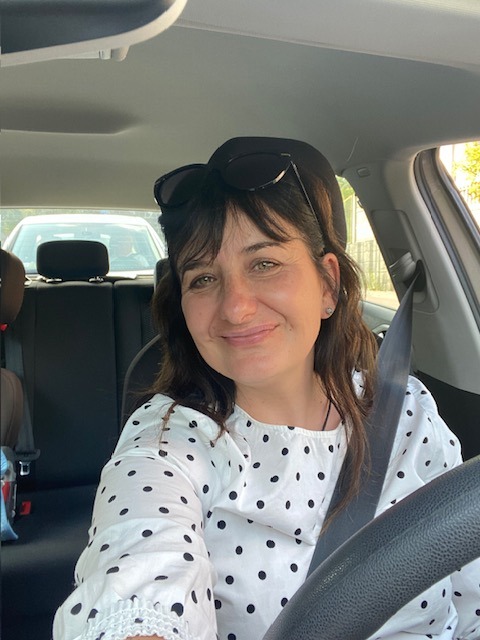 Kobieta z uśmiechem, w samochodzie, zapięta pasami, w bluzce w kropki i okularach na głowie, robi selfie na tle wnętrza samochodu.