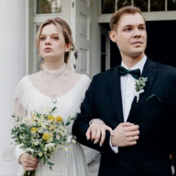 Elegancka para młoda w klasycznych strojach, panna młoda z bukietem białych i żółtych kwiatów, pozuje na tle białych kolumn i drzwi.