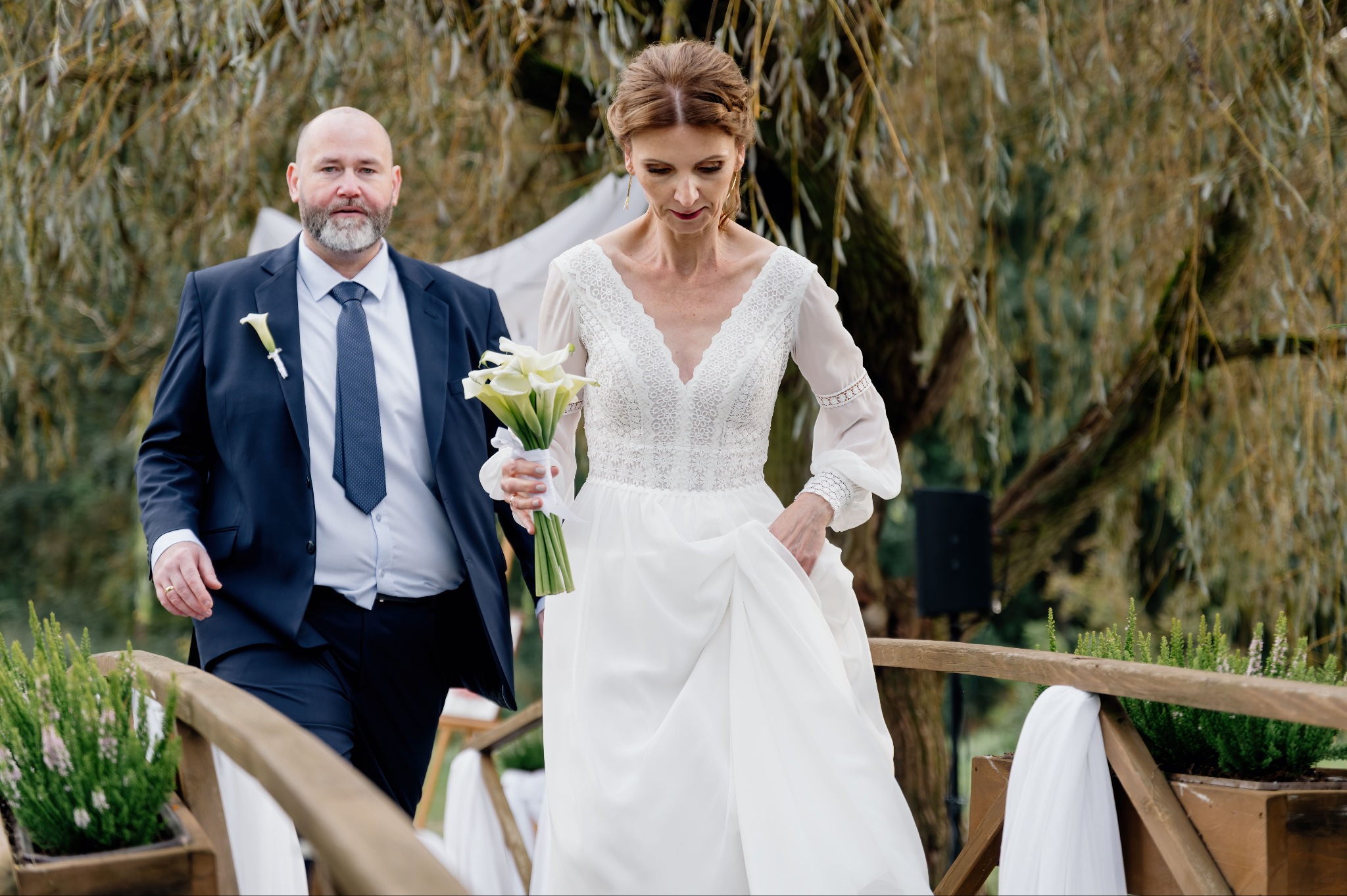 Para młoda przechodzi przez ozdobiony drewniany mostek; panna młoda w koronkowej sukni z bukietem kalii, pan młody w granatowym garniturze z butonierką.