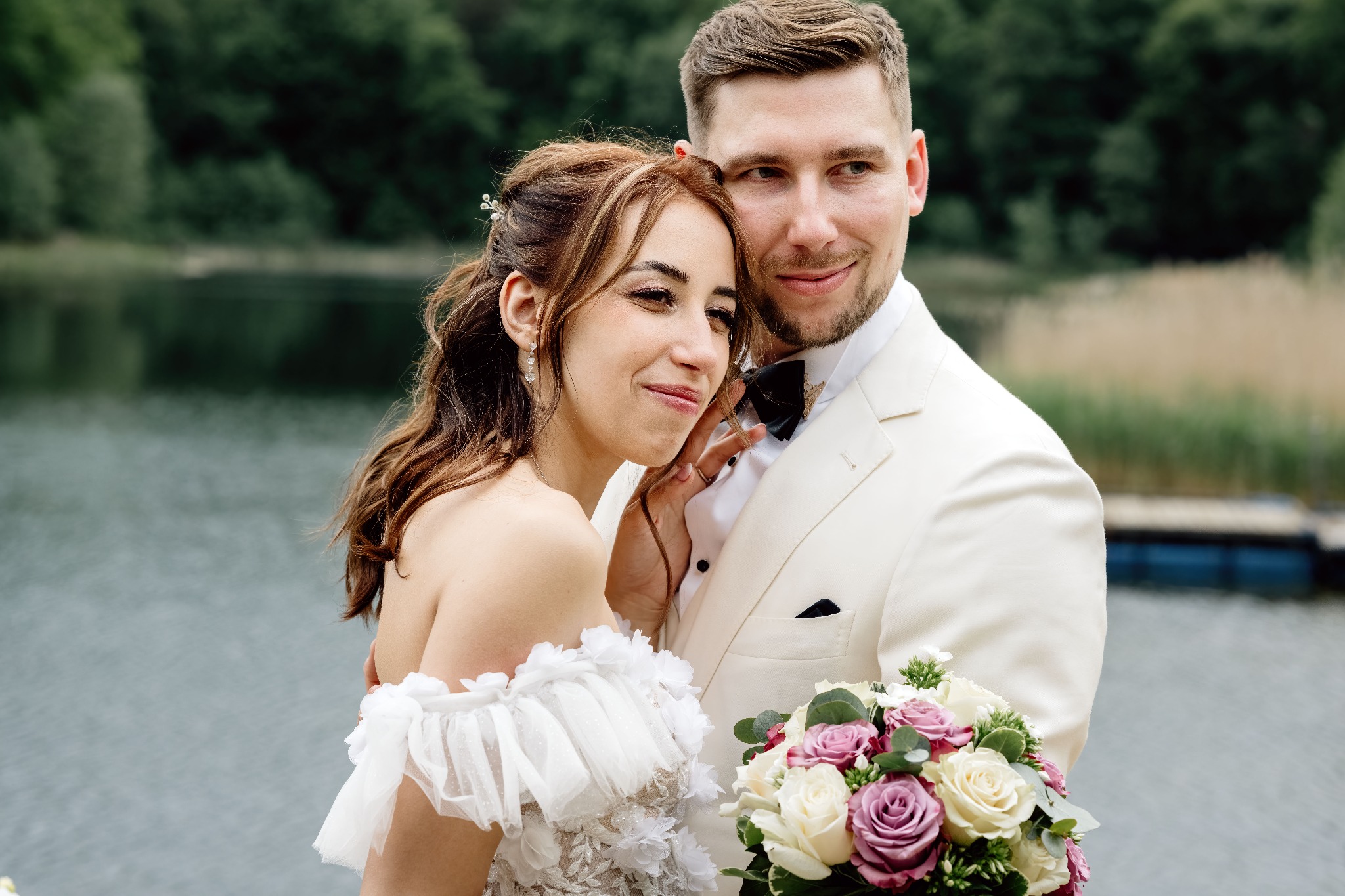 Uśmiechnięta para młoda w białych strojach, on z muchą, ona z bukietem, pozuje na tle jeziora i zieleni. Romantyczna sesja ślubna w plenerze.