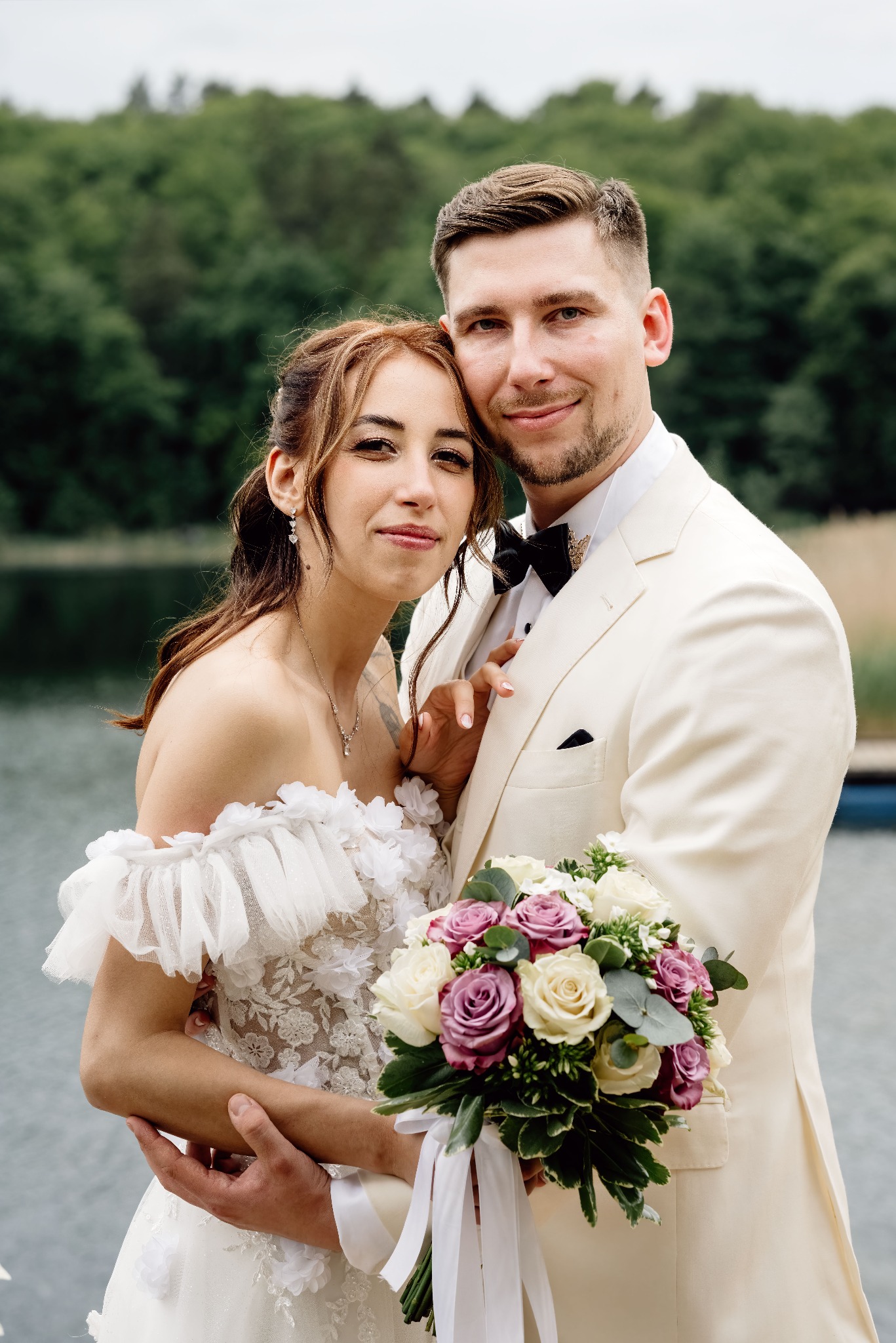 Uśmiechnięta para młoda w plenerze: panna młoda w sukni z kwiatami, pan młody w kremowym smokingu, bukiet róż, jezioro i zieleń w tle. Romantyczna sesja ślubna.