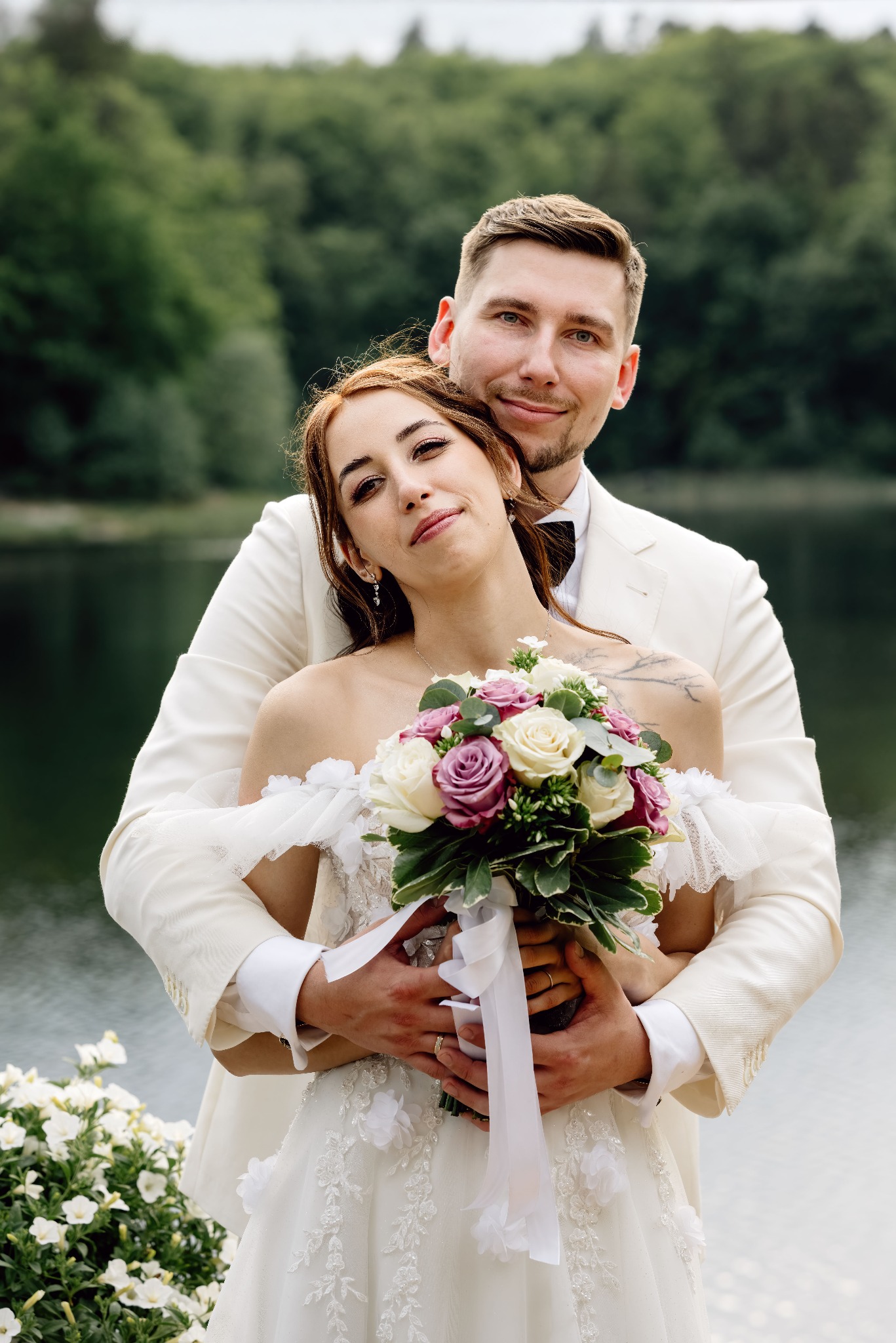 Uśmiechnięta para młoda w bieli, on obejmuje ją od tyłu, trzymając bukiet różowych i białych róż na tle jeziora i zieleni. Delikatny makijaż panny młodej.