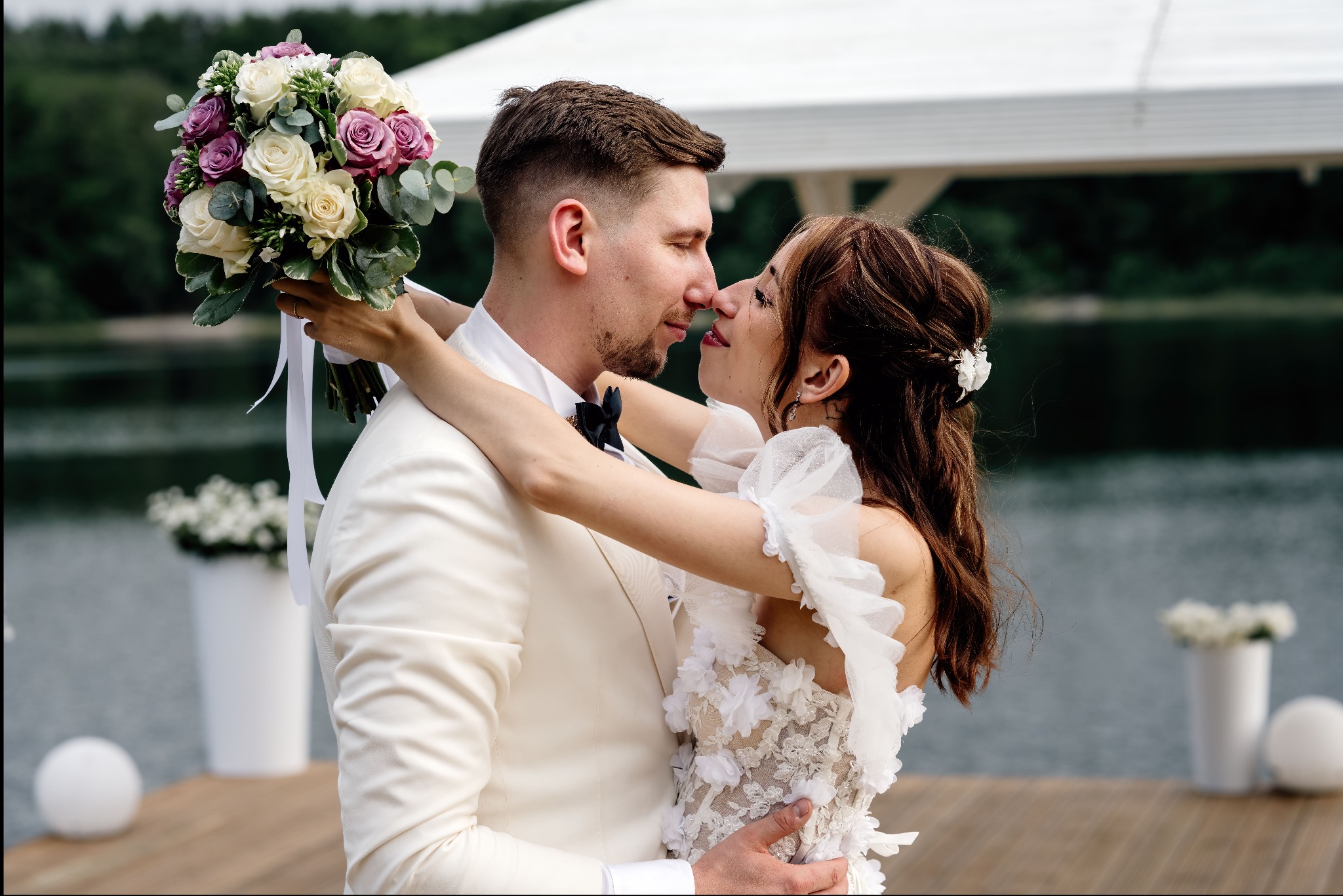 Para młoda w objęciach na tle jeziora. Panna młoda trzyma bukiet z białych i fioletowych róż. Delikatny makijaż panny młodej. Elegancki garnitur pana młodego.