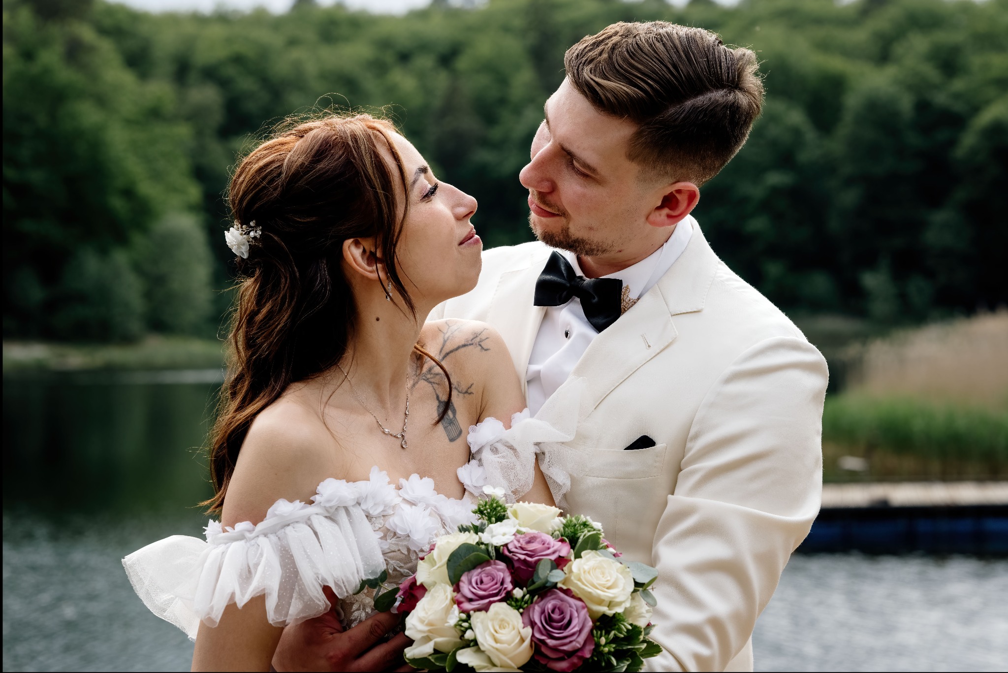 Para młoda w białych strojach, spoglądająca na siebie z czułością, na tle zieleni i jeziora. Bukiet z róż w dłoniach.