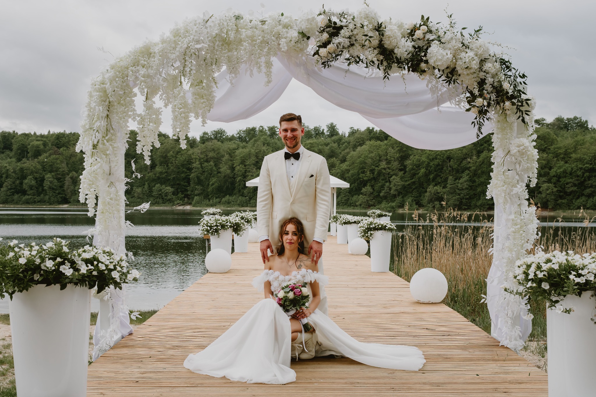 Para młoda na drewnianym pomoście pod dekorowanym białymi kwiatami łukiem, z jeziorem i zielenią w tle. Pan młody w kremowym garniturze, panna młoda w sukni.
