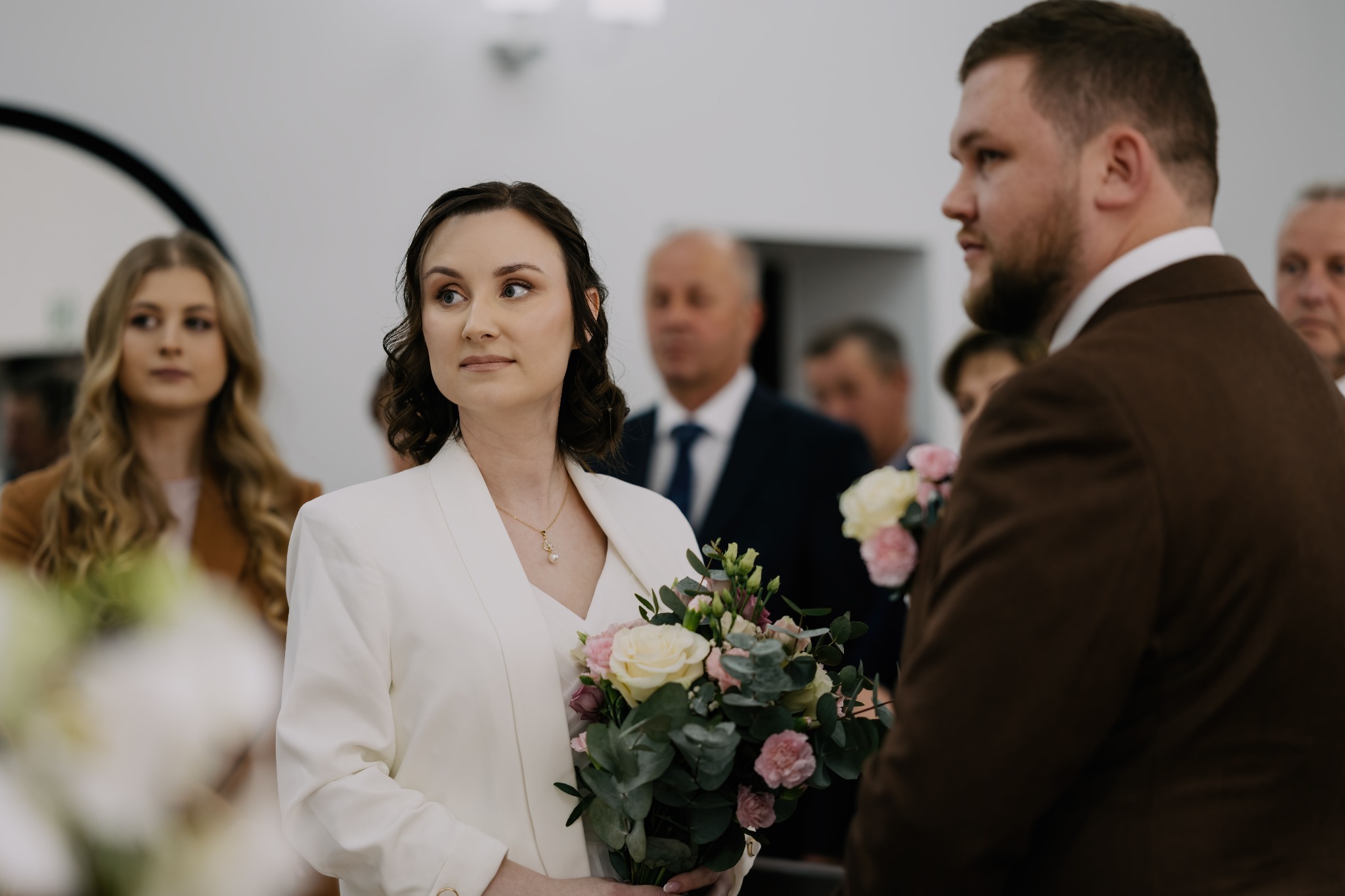 Panna młoda w białym garniturze trzyma bukiet róż i eukaliptusa, spoglądając w bok podczas ceremonii ślubnej. Pan młody w brązowym garniturze obok. W tle goście.