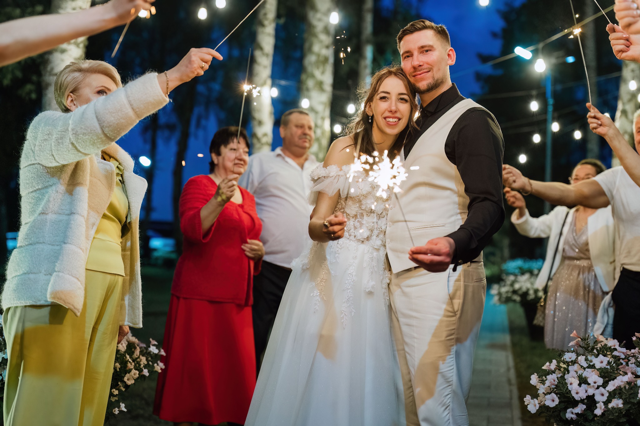 Uśmiechnięta para młoda z iskierkami otoczona gośćmi weselnymi w plenerze, wieczorową porą. W tle drzewa i dekoracyjne oświetlenie, tworzące magiczną atmosferę.