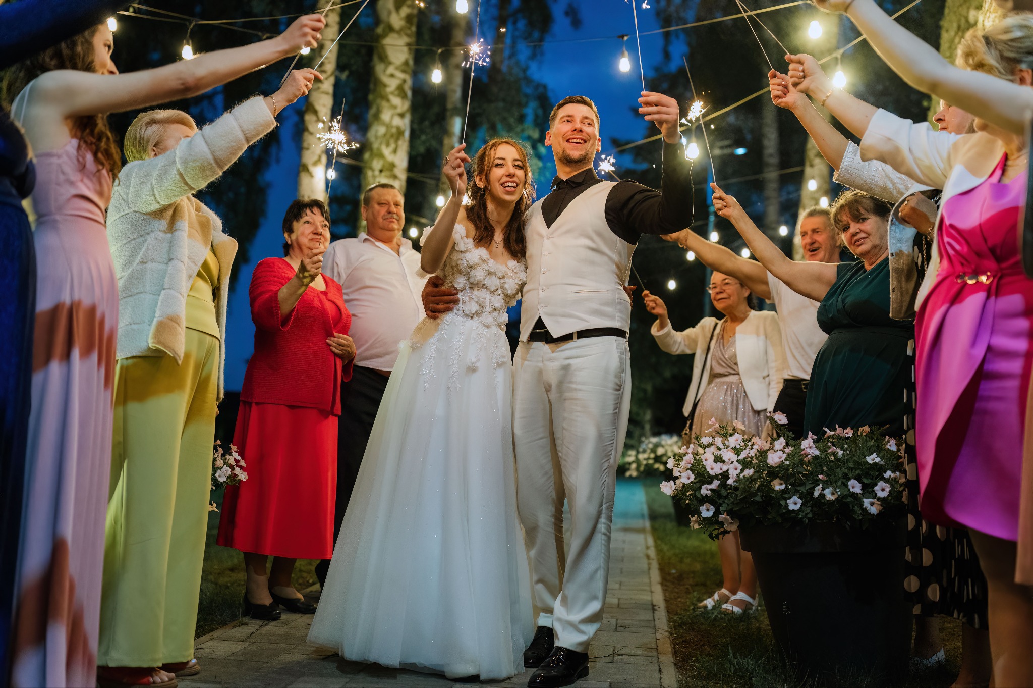 Uśmiechnięta para młoda otoczona gośćmi z zimnymi ogniami, w tle girlandy żarówek i drzewa. Ujęcie z dołu, podkreślające radość i celebrację.