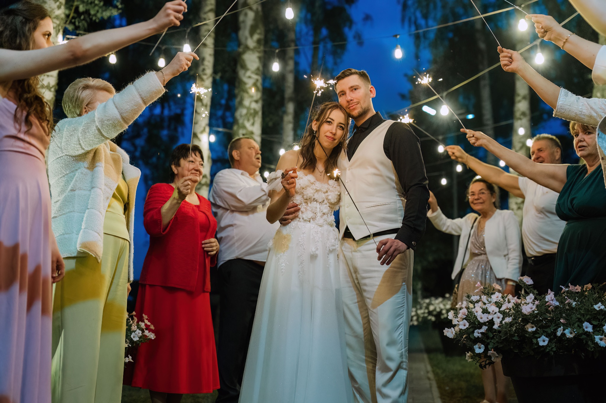 Para młoda otoczona gośćmi z zimnymi ogniami, w tle drzewa i girlandy światełek, uśmiechy, radosna atmosfera podczas uroczystości ślubnej.