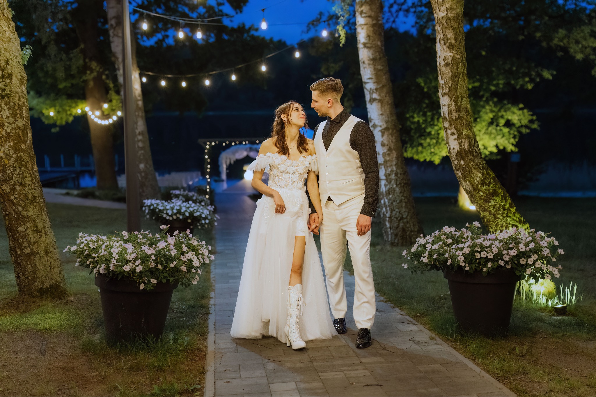 Para młoda w białych strojach na tle dekoracji ślubnych i oświetlenia parkowego. Panna młoda ma nietypowe buty. Ujęcie w nocy, romantyczna atmosfera.