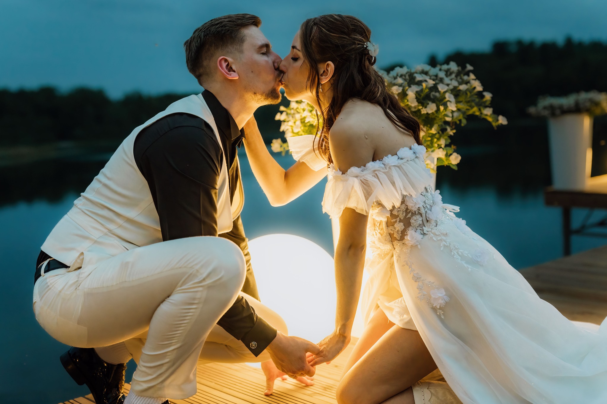 Para młoda w pocałunku na drewnianym pomoście nad wodą, oświetlona ciepłym światłem lampy, z dekoracjami kwiatowymi w tle i spokojnym, wieczornym krajobrazem.