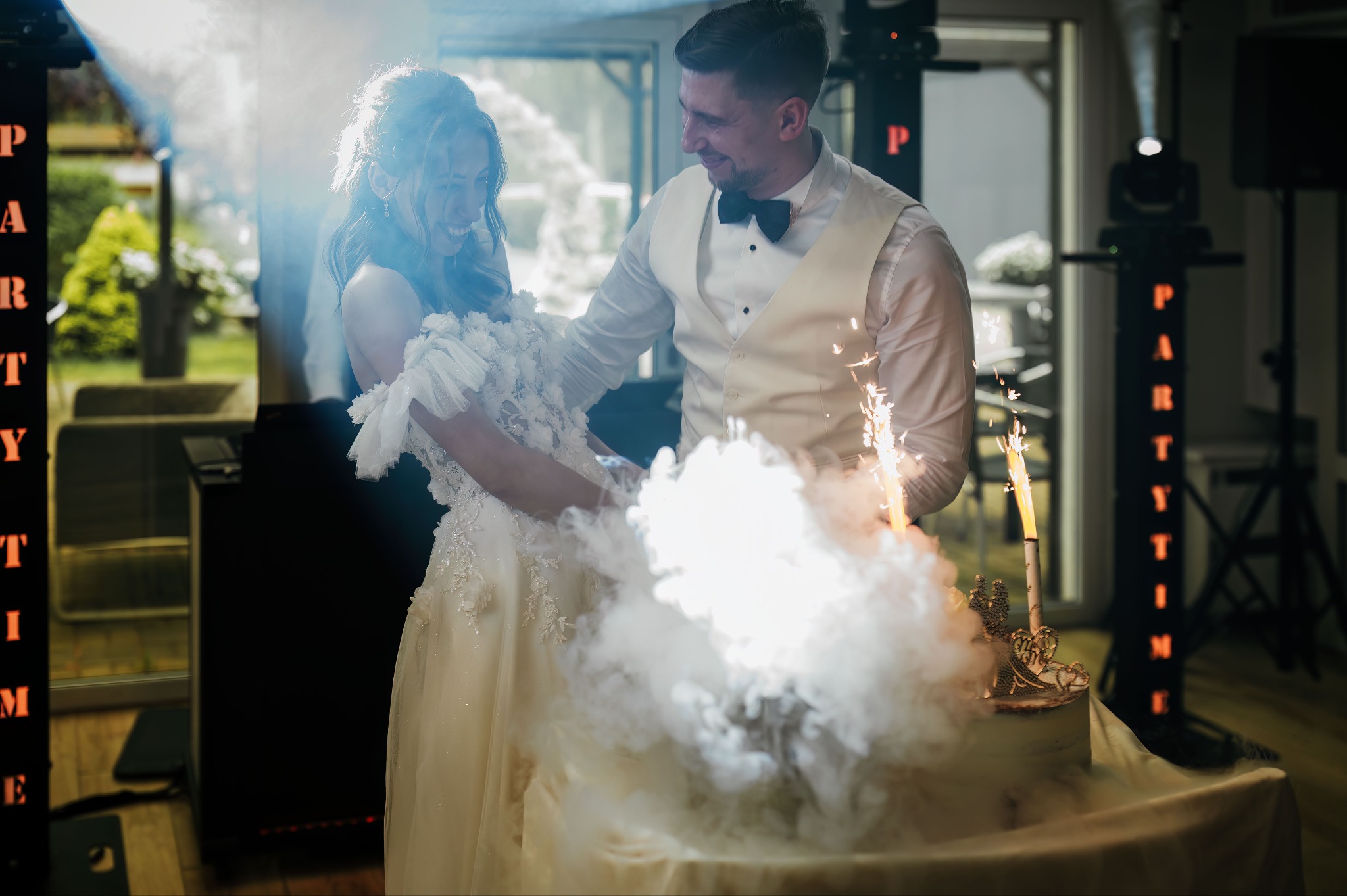 Uśmiechnięta para młoda kroi tort weselny z fontannami iskier na tle dekoracji 'Party Time' i okna z zielenią. Pan młody w muszce i kamizelce.