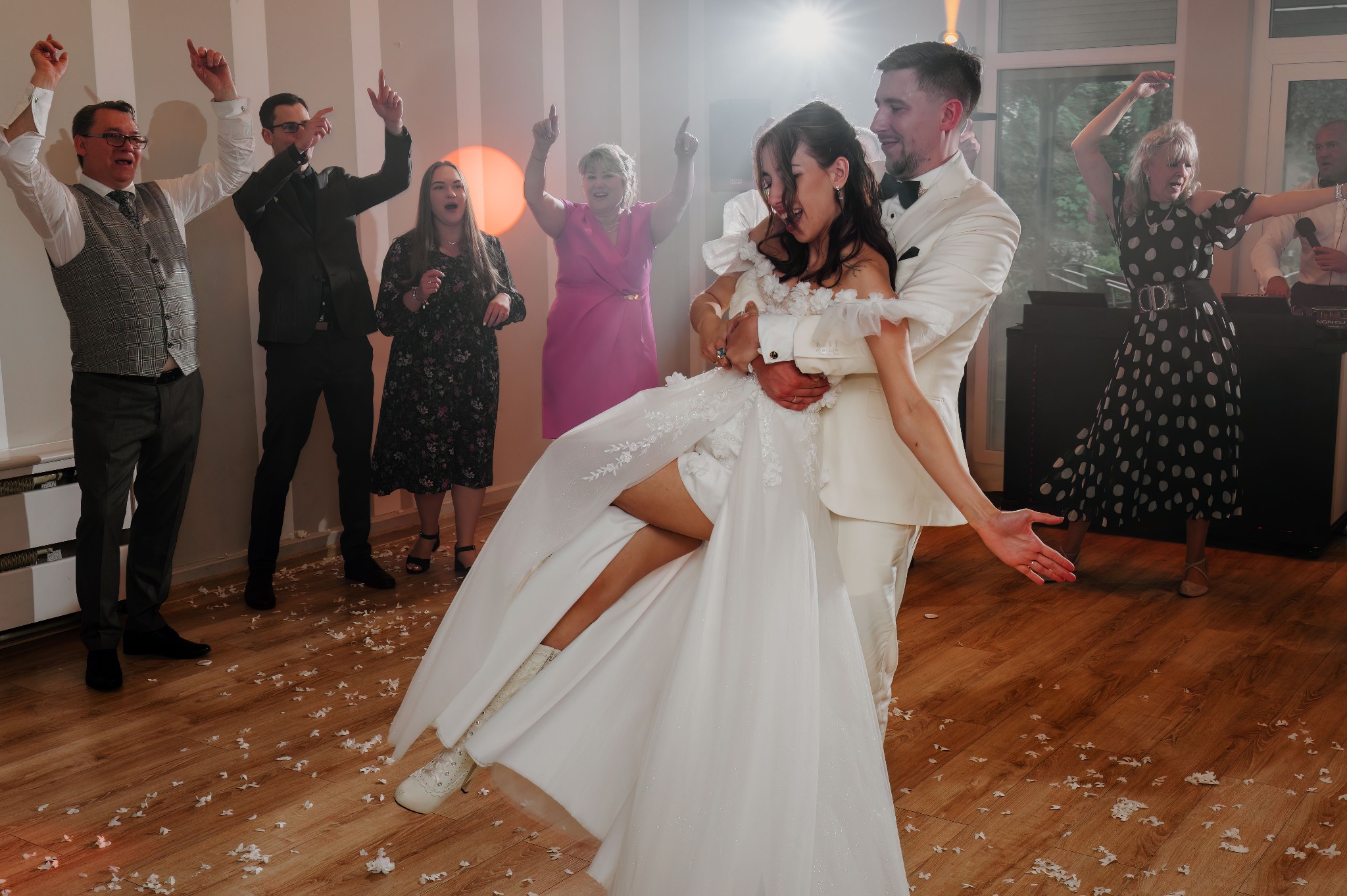 Rozbawiona para młoda w białych strojach tanecznych na parkiecie, otoczona gośćmi weselnymi podczas zabawy. Na podłodze leżą białe płatki kwiatów.