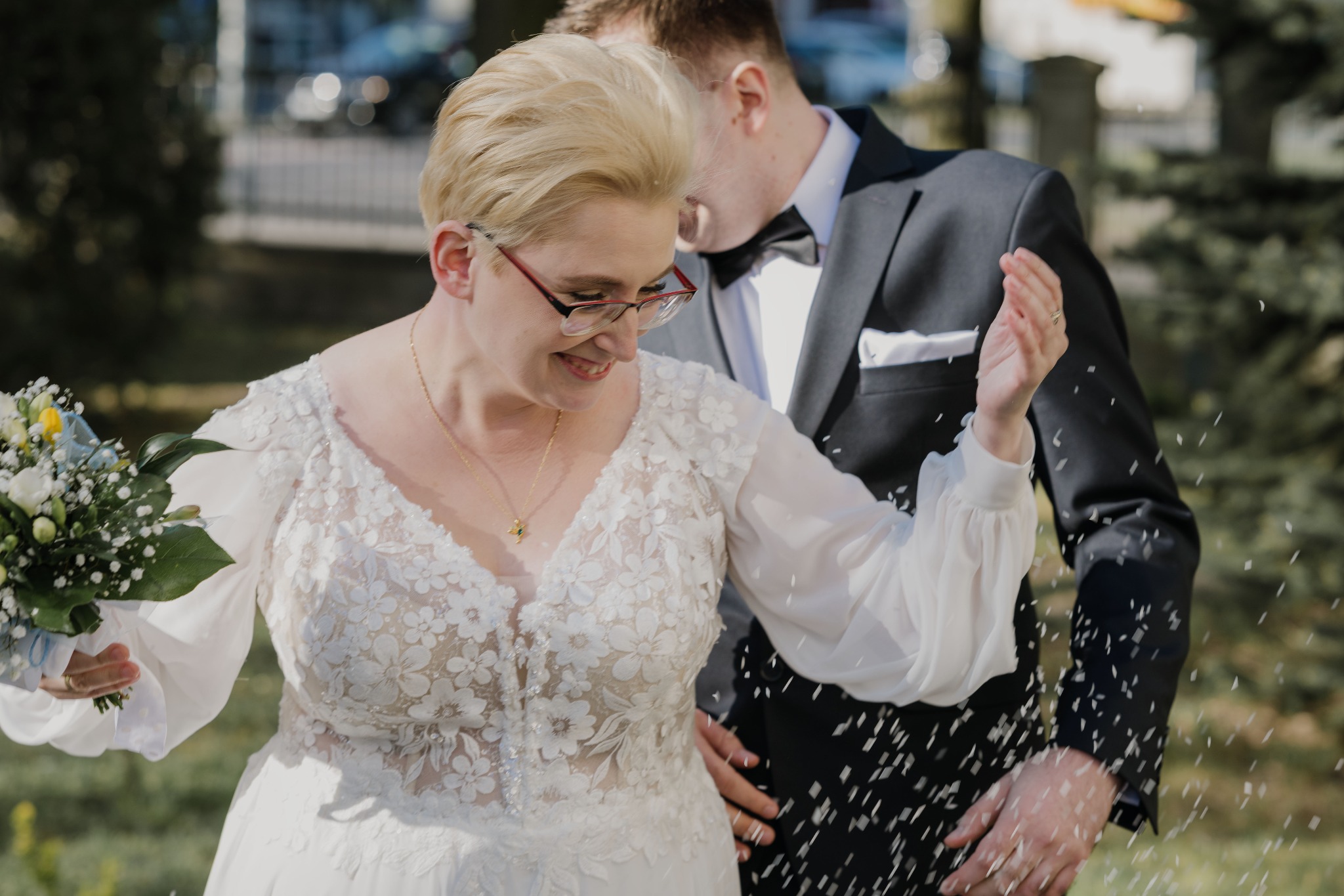 Para młoda w Toruniu podczas ceremonii zaślubin. Panna młoda w sukni z bukietem, obok pan młody w garniturze. W tle zieleń i budynek, konfetti w powietrzu.