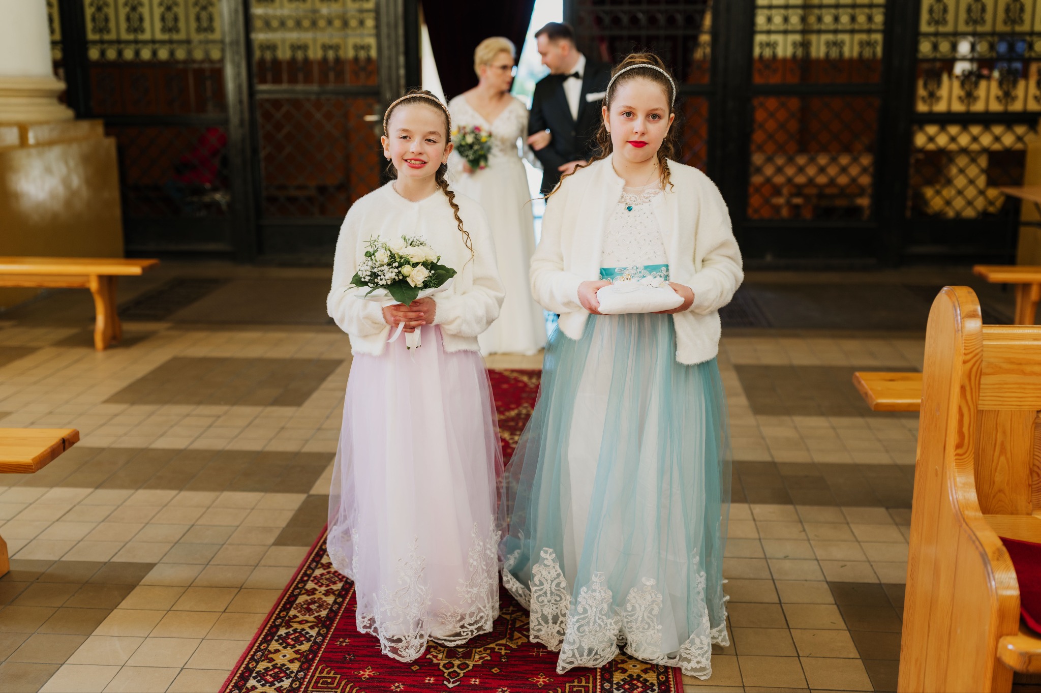 Dwie dziewczynki w sukienkach druhny niosące poduszki z obrączkami i bukiet na tle pary młodej w kościele, uchwycone w ciepłym oświetleniu.