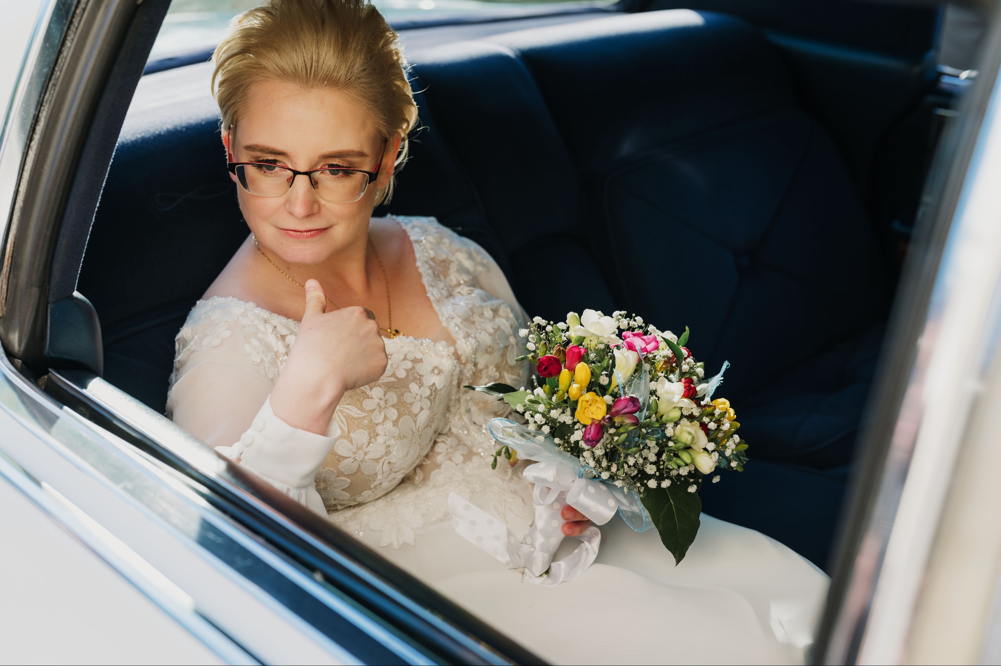 Panna młoda w sukni z koronką i okularach, siedzi w samochodzie, trzyma bukiet ślubny z tulipanami. Ujęcie z wnętrza pojazdu, z otwartymi drzwiami.