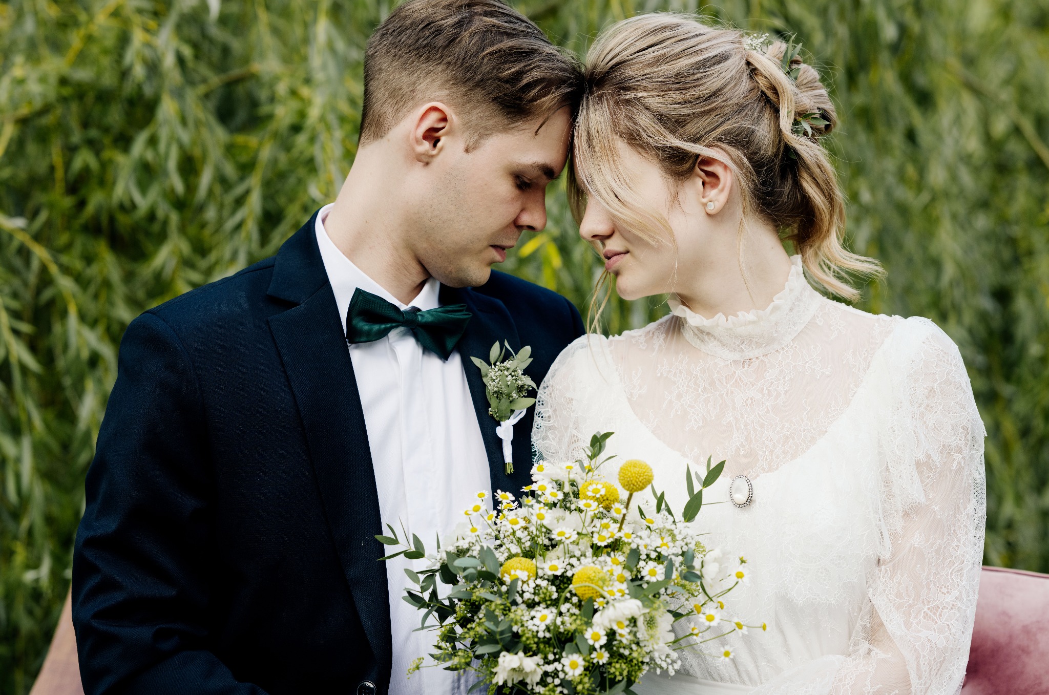 Elegancka para młoda, stykająca się czołami, z bukietem polnych kwiatów na tle zieleni. Pan w granatowym smokingu i zielonej muszce, panna młoda w koronkowej sukni.