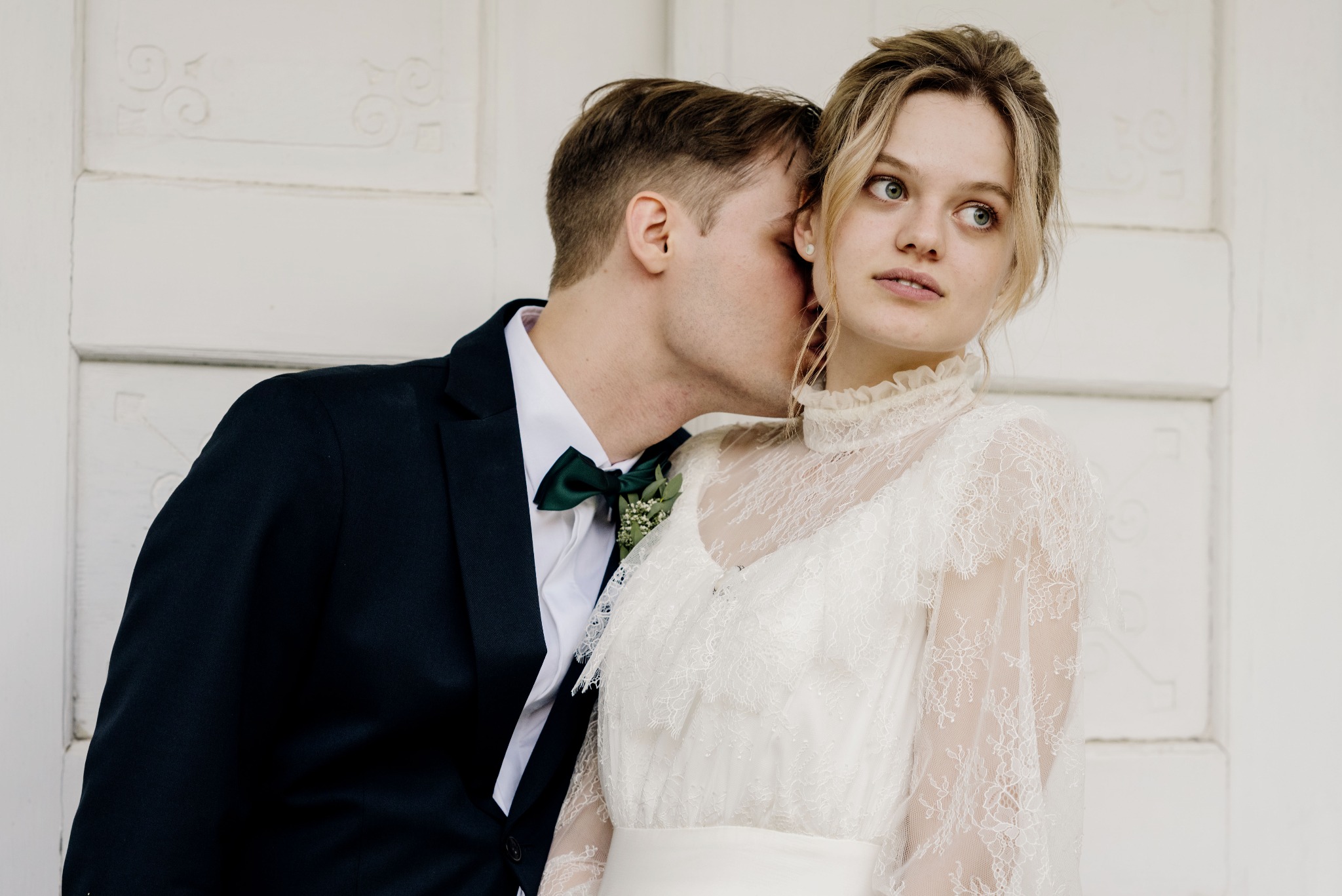 Elegancka para młoda: mężczyzna w granatowym smokingu z zieloną muchą szepta coś do ucha pannie młodej w koronkowej sukni na tle białych drzwi.