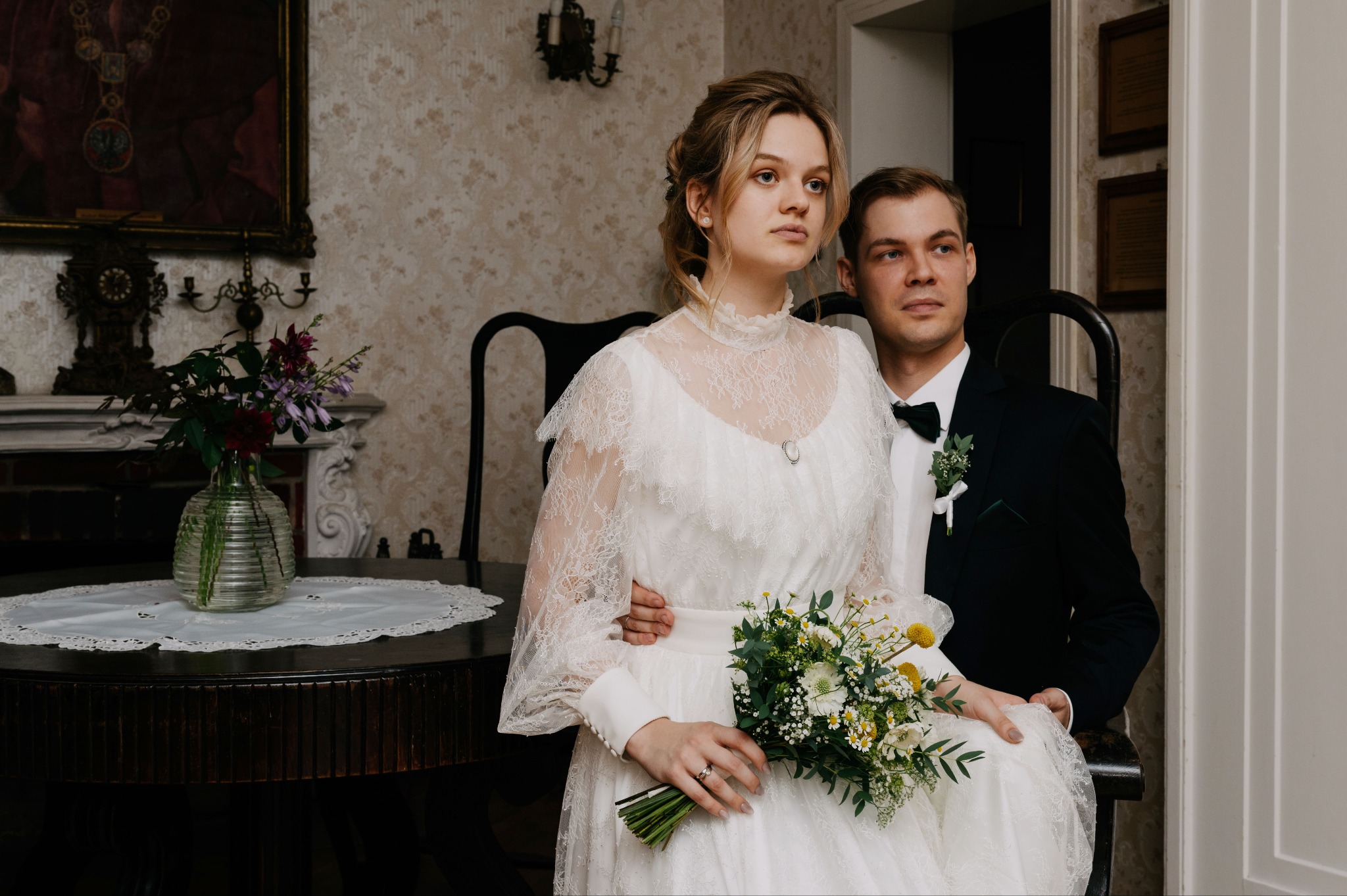 Elegancka para młoda w stylu retro siedzi na krześle. Panna młoda w koronkowej sukni trzyma bukiet, pan młody w smokingu. Klasyczne wnętrze z obrazami i kwiatami.