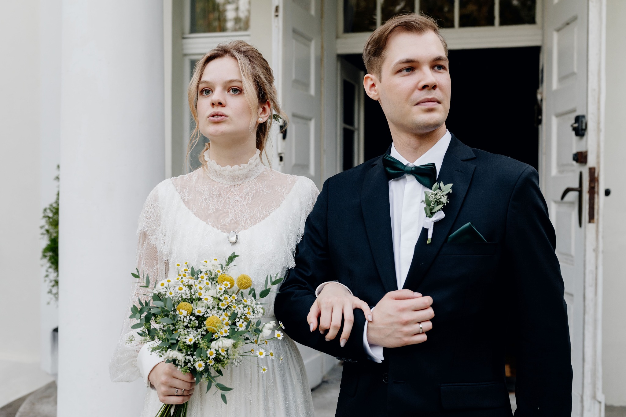 Elegancka para młoda w klasycznych strojach, panna młoda z bukietem białych i żółtych kwiatów, pozuje na tle białych kolumn i drzwi.