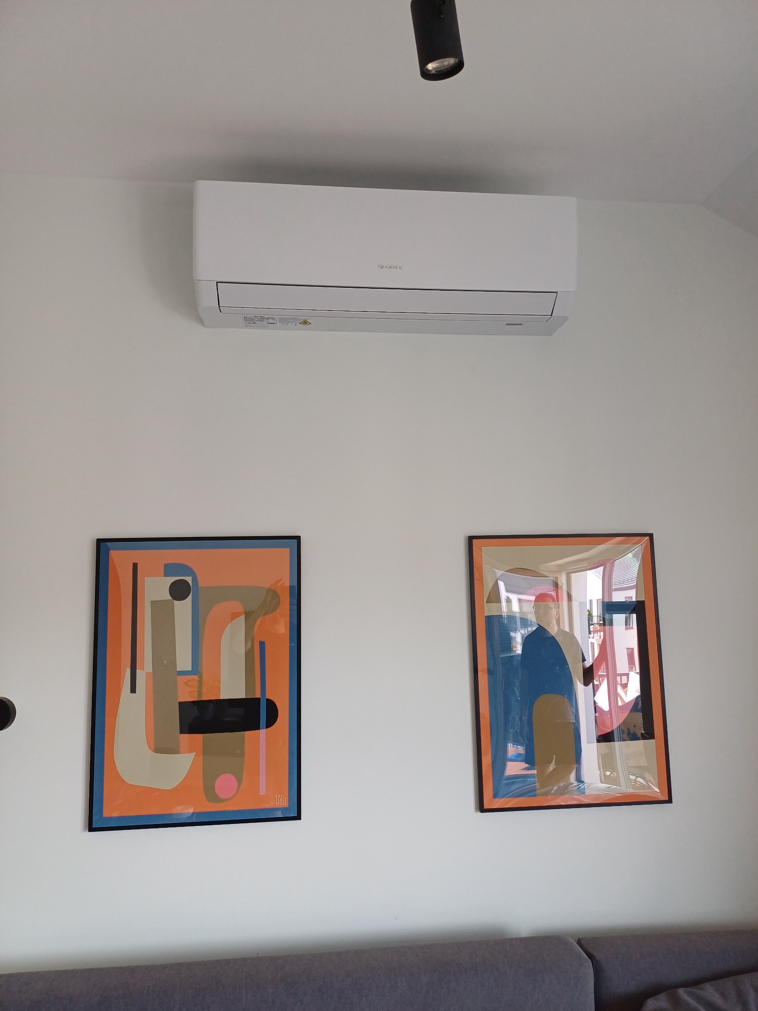 Biały klimatyzator marki Gree zamontowany na ścianie nad dwoma abstrakcyjnymi obrazami w czarnych ramkach i szarą sofą.