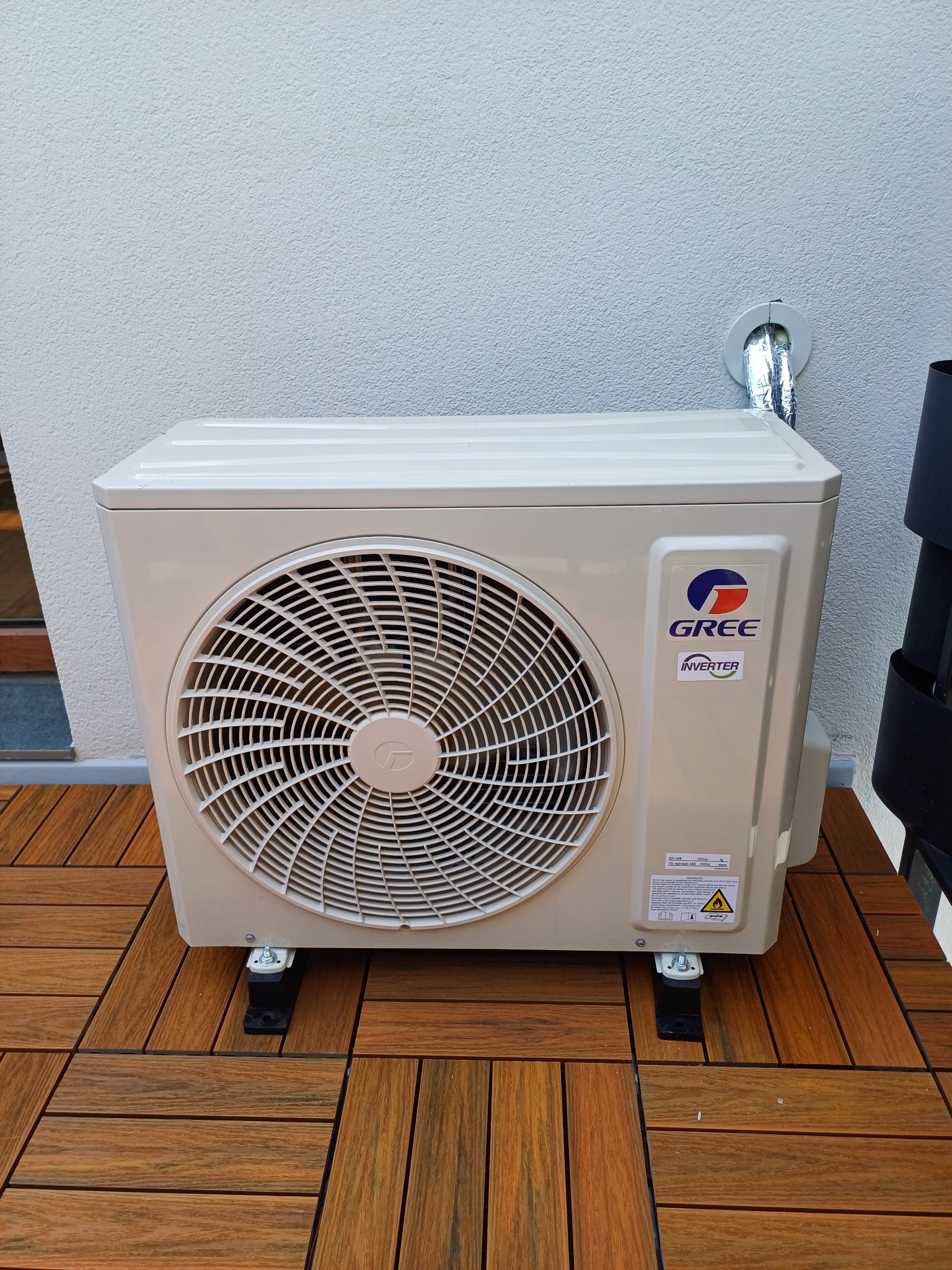 Zewnętrzna jednostka klimatyzacji marki Gree, zamontowana na drewnianym tarasie, z widocznym srebrnym przewodem wentylacyjnym wychodzącym ze ściany.