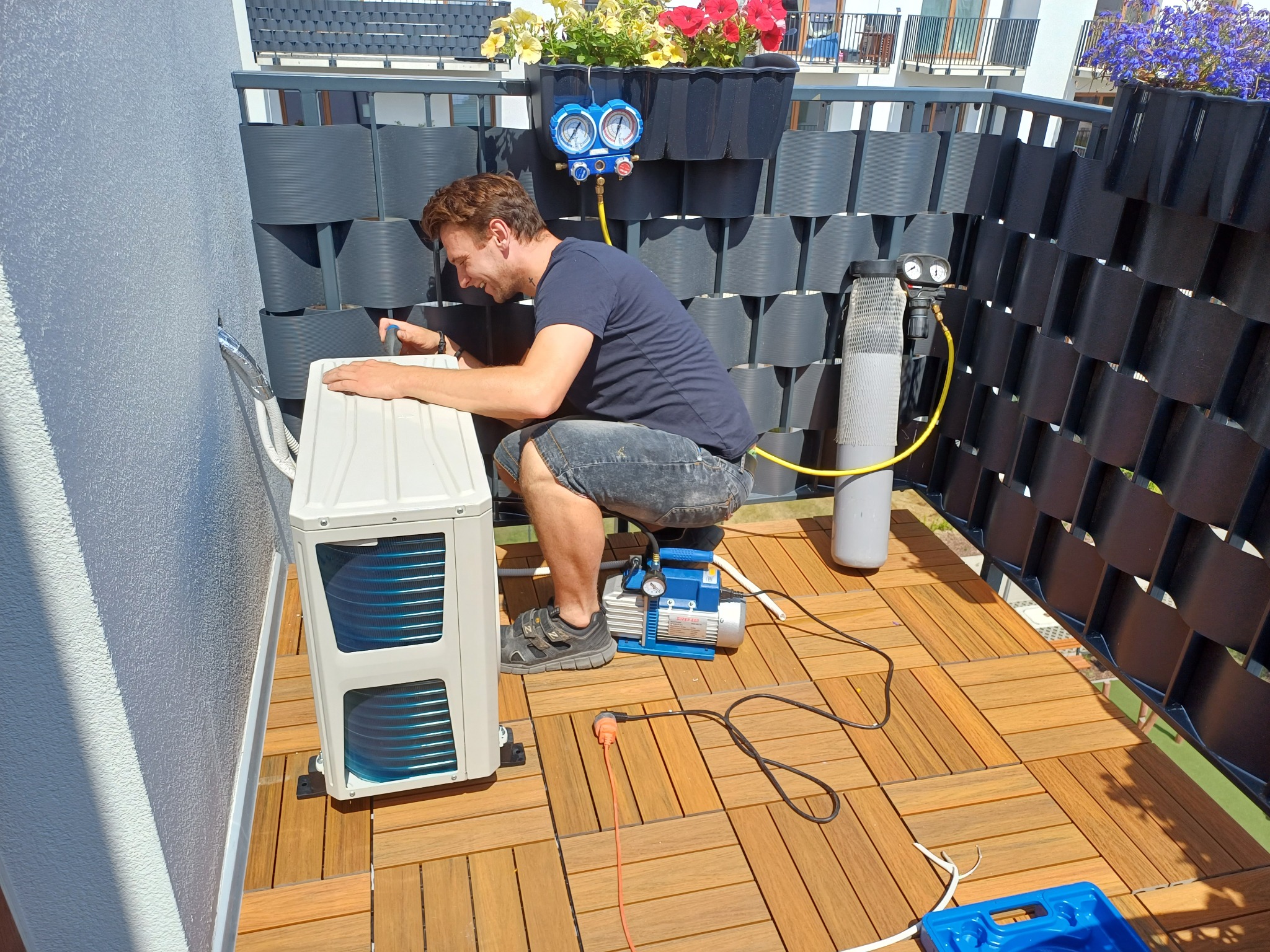 Mężczyzna w trakcie montażu jednostki zewnętrznej klimatyzacji na balkonie, widoczne narzędzia i mierniki, w tle kwiaty w donicach na balustradzie.