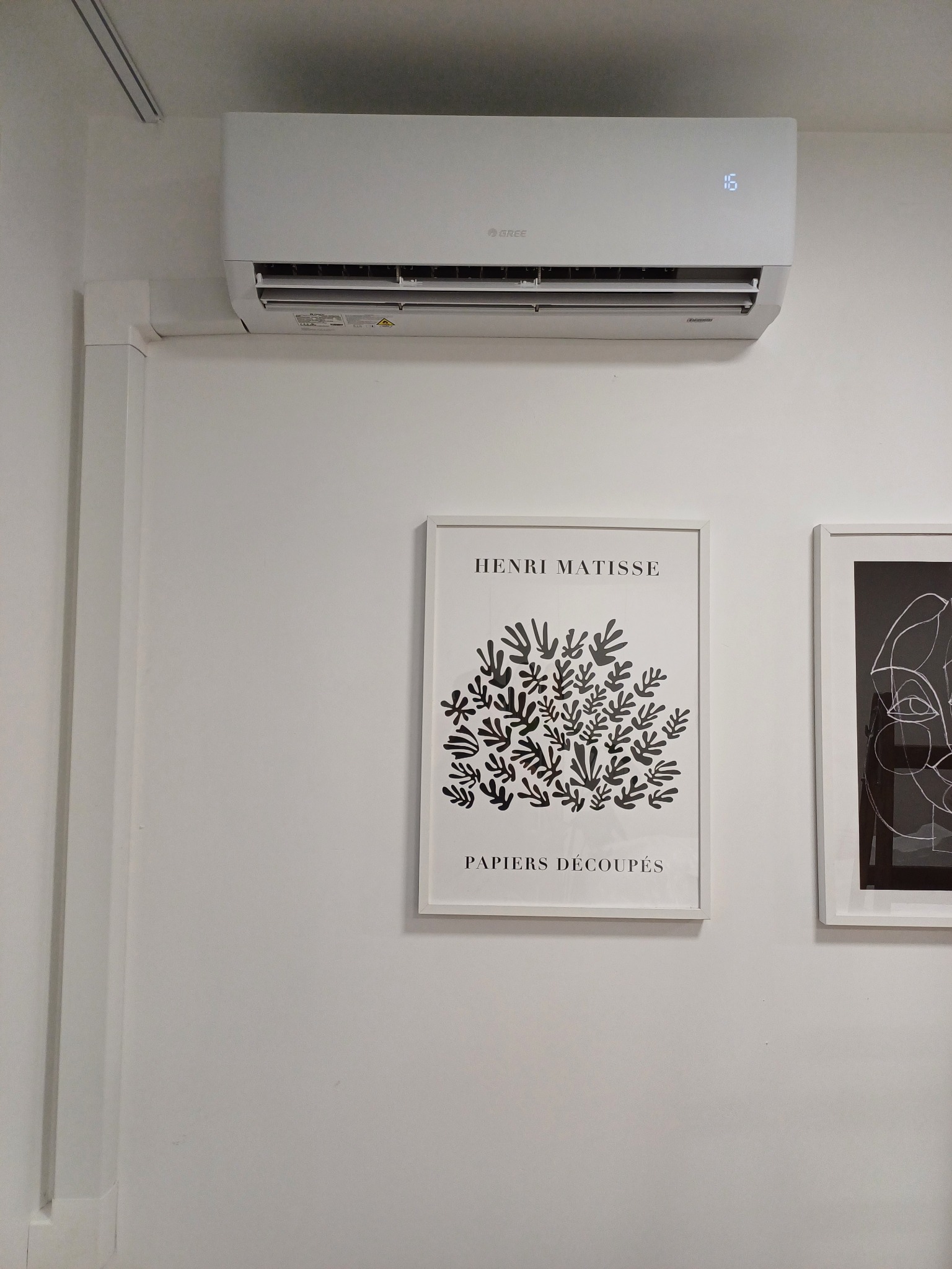Biały klimatyzator marki Gree zamontowany na ścianie, obok wiszą oprawione grafiki Matisse'a w białych ramkach.
