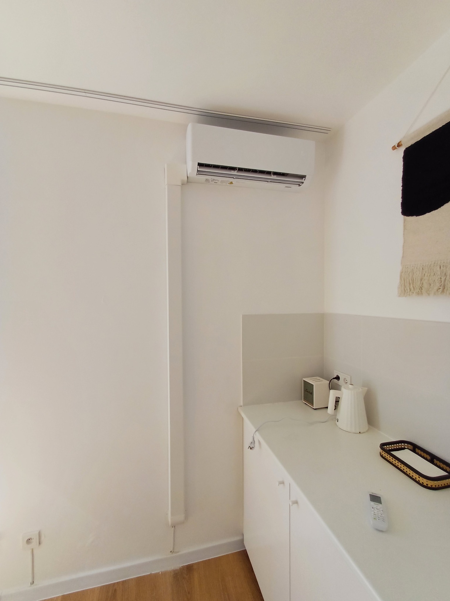 Biały klimatyzator ścienny w minimalistycznym wnętrzu z drewnianą podłogą, białą szafką i dekoracyjnym kilimem na ścianie.