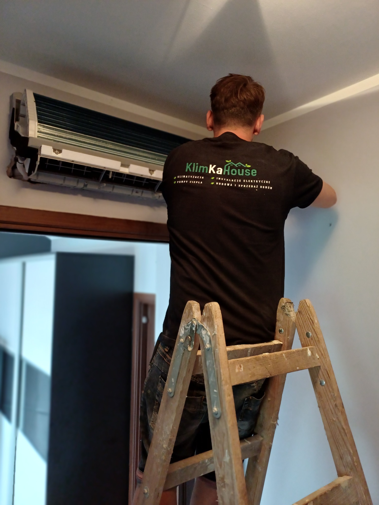 Mężczyzna na drewnianej drabinie montuje klimatyzator ścienny w pomieszczeniu. Na koszulce widoczne logo firmy.